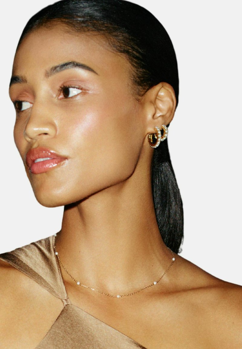 Gouden creolen met heldere accenten, delicate gouden ketting met kleine kralen, en een gladde satijnen bruine top. Close-up van een profiel.