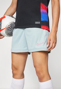 Lichtblauwe sportshorts met een roze Nike-logo, zachte textuur en opgerolde zomen. Speler houdt een zwart-wit patroon voetbal vast.