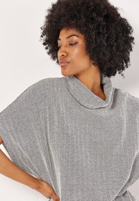 Ylhäältä avonainen poncho-tyylinen paita musta-valkoisessa herringbone-kuosissa, jossa on korkea kaulus ja väljä istuvuus, valmistettu teksturoidusta neulosmateriaalista.