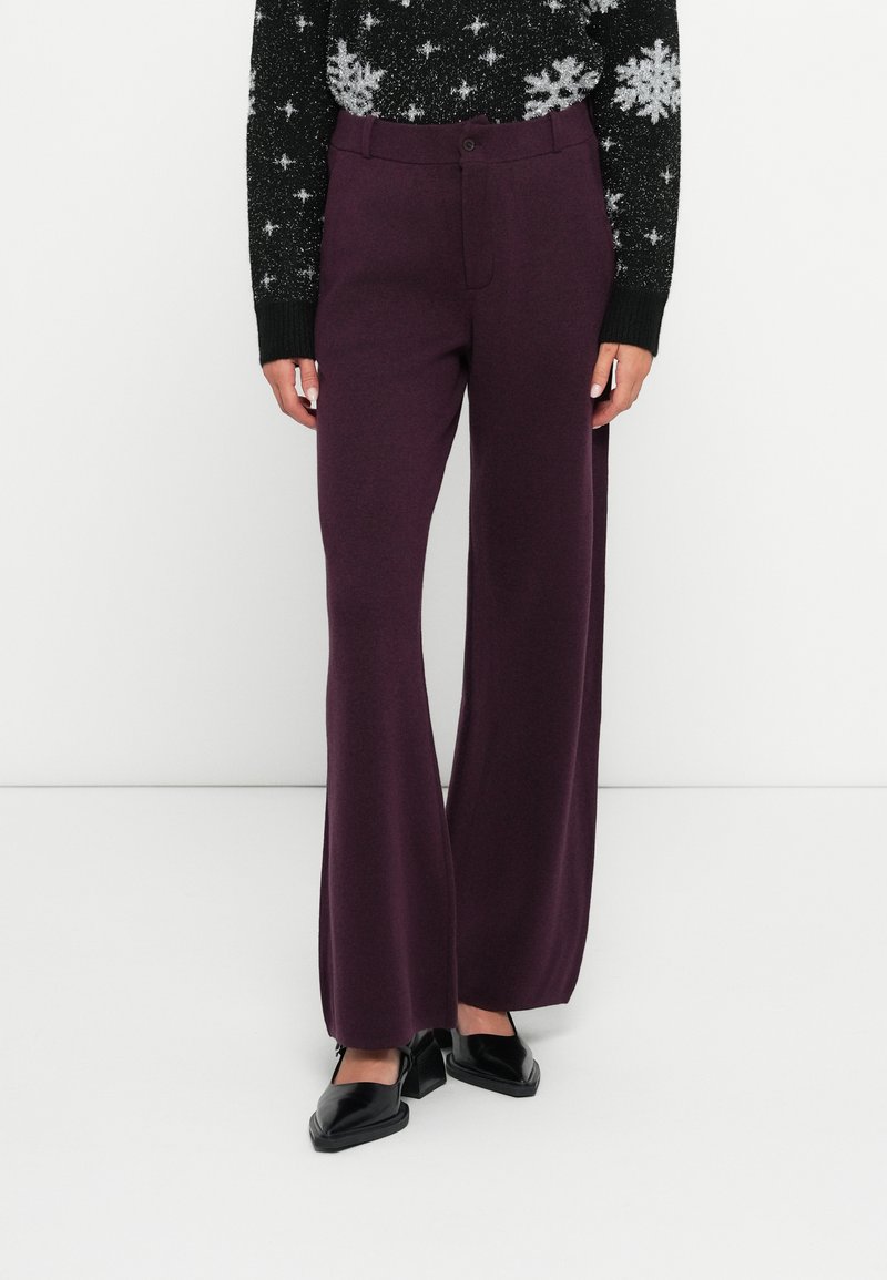 Pantalons larges bordeaux en tissu texturé, avec une fermeture à bouton sur le devant et une taille haute, associés à des chaussures noires.