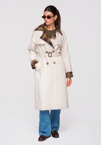 Femme portant un trench-coat beige ceinturé avec col et poignets à carreaux, un haut vert, un jean bleu, des chaussures marron et des lunettes de soleil, posant les mains dans les poches.