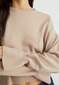 Nainen, joka on pukeutunut beigeen pitkähihaiseen neulepaitaan, jossa on ribbineuleet hihansuissa ja helman alueella, yhdistettynä sinisiin farkkuihin, seinää vasten neutraalissa taustassa.