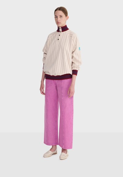 Femme debout portant un pull crème à fines rayures avec col et poignets rouges et marine, pantalon rose large et mocassins tissés beige sur fond uni.