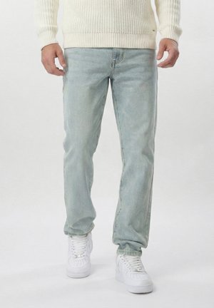 Mężczyzna w jasnoniebieskich dżinsach, białym dzianinowym swetrze i białych trampkach stoi na jednolitym tle.