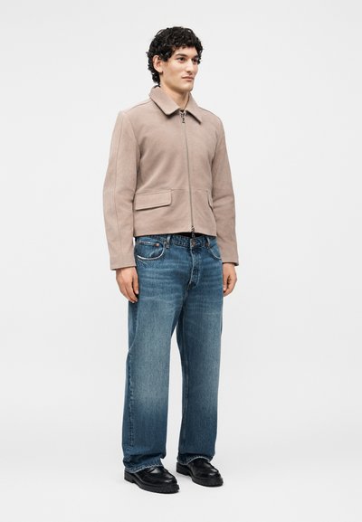 Άνδρας όρθιος, φοράει μπεζ τζάκετ με φερμουάρ, μπλε τζιν και μαύρα παπούτσια σε απλό λευκό φόντο.