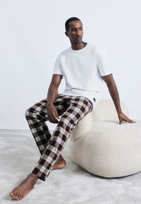 Άνδρας με λευκό μπλουζάκι και καφέ καρό παντελόνι καθισμένος ξυπόλητος σε μια χαμηλή, στρογγυλή μπεζ καρέκλα πάνω σε ένα ανοιχτό χρωματισμένο χαλί.