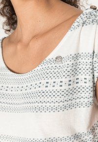 Bílá košile s modrými geometrickými vzory, kulatým výstřihem a krátkými rukávy. Obsahuje malé kovové logo na výstřihu.