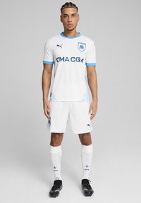 Valge spordijersey siniste aktsentidega, ümmarguse kaelusega, lühikeste varrukatega ja logoga. Koos valgete lühikeste pükste ja sokkidega ning mustade spordijalatsitega.