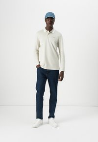 Camisa polo de manga larga color crema, gorra azul, vaqueros de mezclilla oscuros y zapatillas blancas. La polo presenta un cuello blanco en contraste y detalle de logo.
