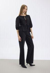 Svart peplum-bluse med puffede ærmer og snøredetalje, kombinert med svarte vide jeans og spisse hæler, på en lys bakgrunn.