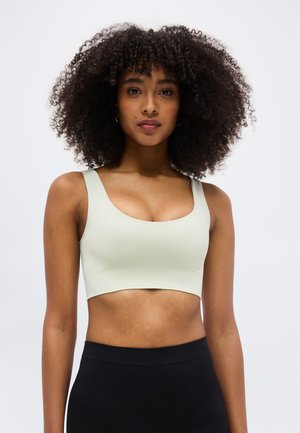 Kvinde med krøllet hår iført en lys grøn cropped tanktop og højtaljede sorte bukser, står foran en ensfarvet lys baggrund.