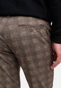 Primer plano de una persona que lleva pantalones marrones estampados con un bolsillo lateral y detalle de botón, junto con un suéter negro.