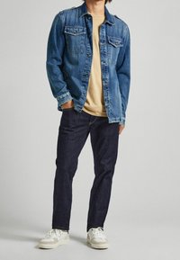 Ljusblå jeansjacka med två bröstfickor, beige t-shirt under, mörkblå jeans och vita sneakers med ljusa detaljer.