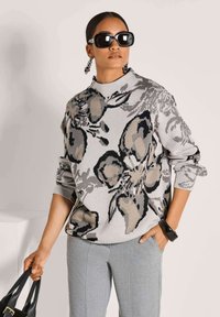 Frau trägt einen übergroßen Pullover mit floralem Muster, graue Hosen, große schwarze Sonnenbrille, auffällige Ohrringe, eine schwarze Uhr und hält eine schwarze Handtasche.