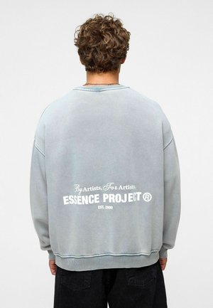 Vervaagde blauwe sweatshirt met een ronde hals en geribbelde manchetten, met op de achterkant witte tekst: "ESSENCE PROJECT EST. 2000."