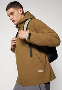 Veste imperméable marron avec capuche, fermeture éclair à l'avant et poches latérales. Dotée d'accents gris et d'un patch logo à l'ourlet. Sac à dos porté.