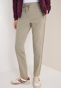 Person mit beigen Kordelzug-Hosen mit seitlichem Streifen, weißen Sneakers mit kastanienbraunen Akzenten und einer kastanienbraunen Zip-Jacke.