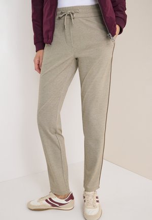 Personne portant un pantalon beige avec cordon de serrage et une bande sur le côté, des baskets blanches avec des accents bordeaux, et une veste bordeaux zippée.