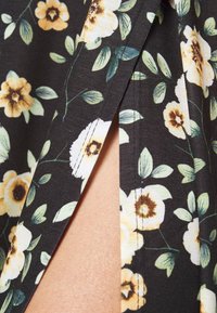 Tissu noir avec imprimé floral jaune et blanc avec des feuilles vertes, montrant une fente en gros plan révélant la peau en dessous.