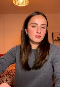 Ung kvinde med langt brunt hår og grå sweater, der sidder indendørs under varmt lys og kigger let nedad og til siden.