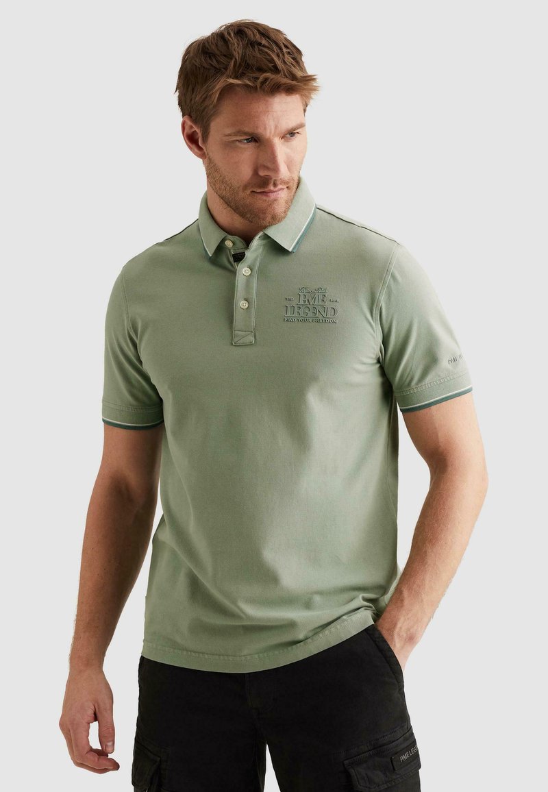 Homme portant un polo vert sauge avec col boutonné et texte brodé, associé à un pantalon cargo noir, regardant sur le côté.
