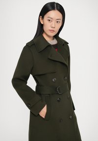 Olijfgroene trenchcoat van een wolmix met een dubbelgeknoopte sluiting, een brede kraag, een ceintuur in de taille en zijzakken met zwarte knopen.