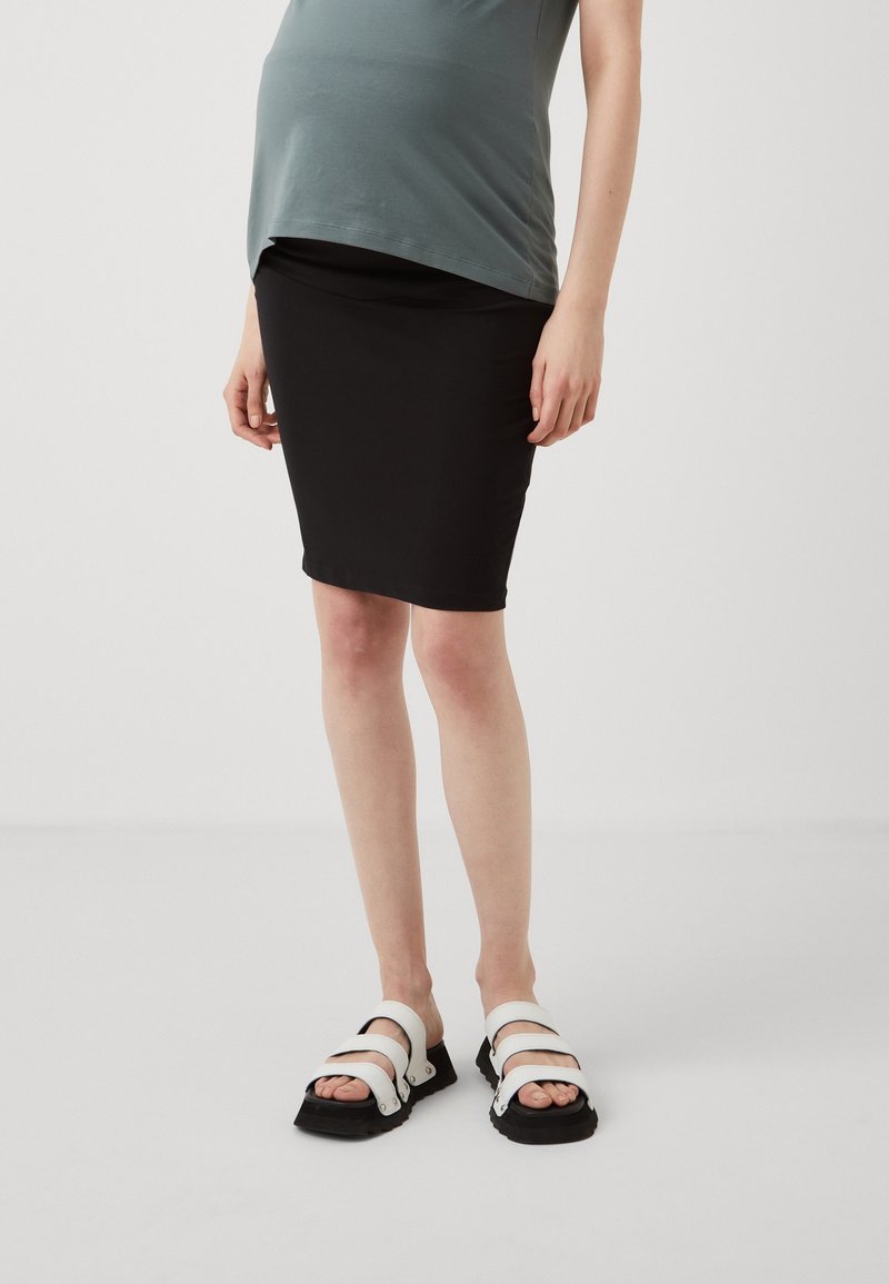 Personne enceinte portant un haut gris, une jupe noire au genou et des sandales blanches, debout sur un fond clair uni.