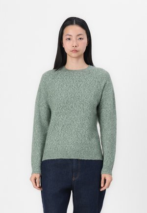 Un pull en tricot vert avec un col rond, des manches raglan, un ourlet côtelé et une finition texturée, associé à un pantalon en denim bleu foncé.