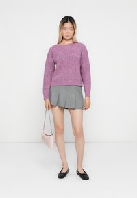 Maglione rosa a maglia con maniche lunghe, abbinato a una gonna grigia plissettata. Tiene in mano una piccola borsa rosa e indossa delle ballerine nere.