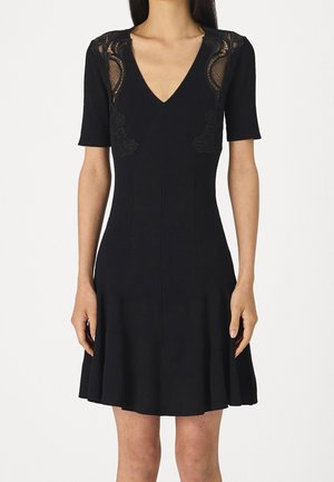 Zwarte jurk met korte mouwen en V-hals met kanten details op de schouders, getailleerde taille en een uitlopende rok die boven de knieën reikt.