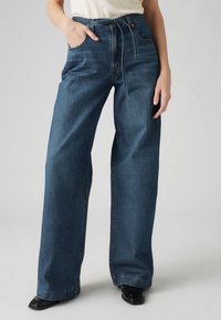 Jeans med vida ben i mörkblå denim med hög midja, justerbar dragsko och framfickor. Slät textur med minimal slitage.