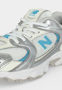 Άσπρο και ασημί παπούτσι για τρέξιμο New Balance με ανοιχτό μπλε λεπτομέρειες, δεμένα κορδόνια και υφασμάτινο επάνω μέρος με υφή.