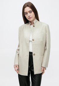 Γυναίκα με ίσια καστανά μαλλιά φοράει μπεζ σακάκι με κουμπιά πάνω από λευκό τοπ και μαύρο δερμάτινο παντελόνι, στέκεται σε απλό λευκό φόντο.