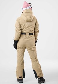 Completo da sci beige con cappuccio, accenti neri sui polsini e orli, dettaglio con cintura in vita, abbinato a guanti neri e a un berretto rosa.