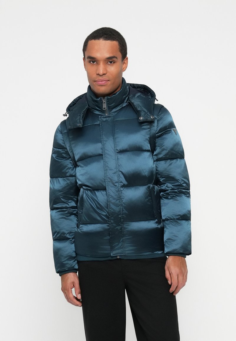 Hombre vistiendo una chaqueta acolchada brillante de color teal con capucha, de pie contra un fondo blanco liso con las manos relajadas a los lados.