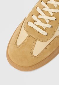 Tan suede sneaker with cream accents, textured upper, flat cotton laces, and rubber sole. Features stitched detailing and scalloped edges.