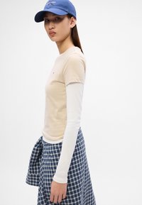 Beige kortermet T-skjorte lagdelt over en hvit langermet topp, parret med et blått rutete skjørt. Blå caps fullfører antrekket.