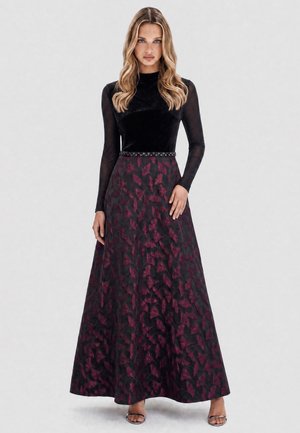 Woman in a long black dress with sheer sleeves and a dark red patterned skirt, standing with one hand resting on her hip.