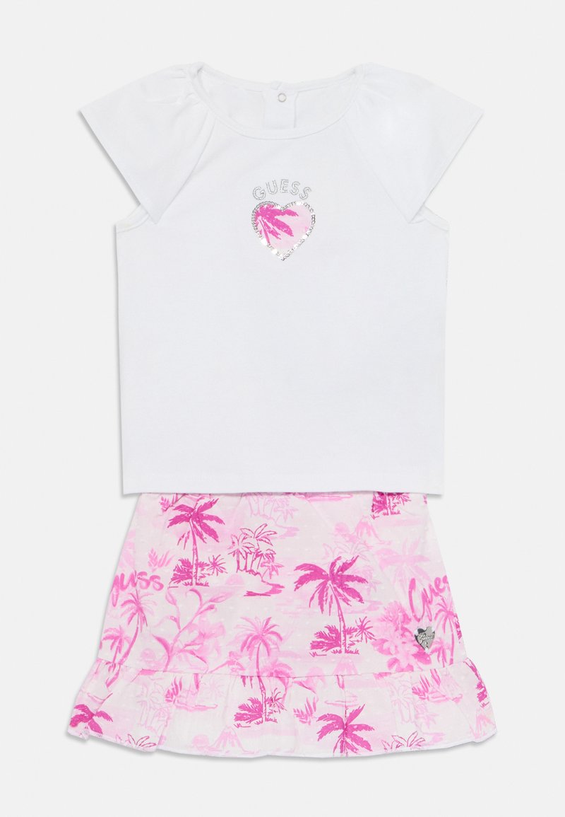 White short-sleeve top with silver "GUESS" logo and pink heart. Pink skirt features palm tree print and ruffled hem, with "GUESS" text.