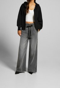 Una donna indossa un hoodie nero con zip, una canotta bianca corta, pantaloni grigi a gamba larga con coulisse e scarpe nere a punta su uno sfondo neutro.