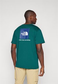 Žalia medvilninė marškinė su mėlynu kvadratiniu grafiniu pavidalu ant nugaros, kuriame užrašyta "THE NORTH FACE" ir frazė "NEVER STOP EXPLORING". Trumpomis rankovėmis.