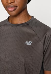 Σκούρο γκρι αθλητικό πουκάμισο από διαπνέον ύφασμα με μικρές οπές. Διαθέτει ένα διακριτικό λογότυπο New Balance σε ανοιχτό γκρι στο στήθος.