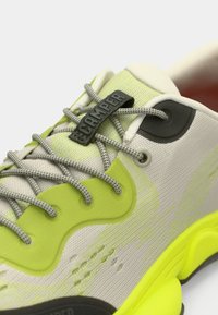 Κοντινό πλάνο ενός ανοιχτού πράσινου και μπεζ αθλητικού παπουτσιού με γκρίζες κορδόνες και μια μαύρη ετικέτα "CAMPER" στη γλώσσα, με υφασμάτινη υφή και ζωντανή σόλα.