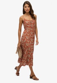 Luźna, rdzawa midi sukienka w wzór paisley, na cienkich ramiączkach, z guzikami i lekkiego materiału, połączona z brązowymi sandałami na obcasie.