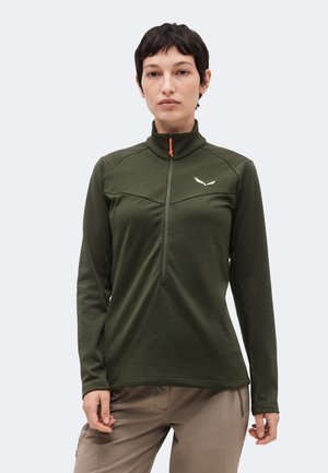 Mujer con cabello corto y oscuro, con una sudadera con cremallera de manga larga de color verde oliva y pantalones beige, de pie frente a un fondo claro y liso.