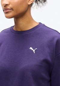 Άτομο που φοράει μοβ φούτερ με λαιμόκοψη κανονική και μικρό λευκό λογότυπο Puma στο αριστερό στήθος, εμφανιζόμενο από τον λαιμό έως το επάνω μέρος του κορμού.