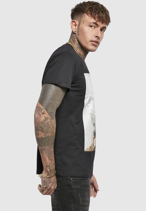 Mężczyzna z tatuażami na szyi, twarzy i ramionach, ubrany w czarną koszulkę z nadrukiem i ciemne dżinsy, stojący bokiem na jednolitym tle.