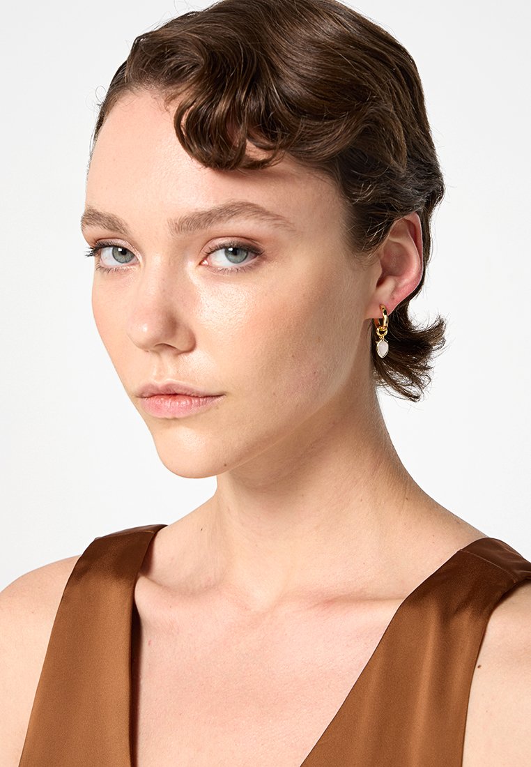 Złote kolczyki w kształcie obręczy z akcentem w postaci perły w kształcie kropli, umieszczone na gładkiej, jasnej karnacji z neutralnym tłem.