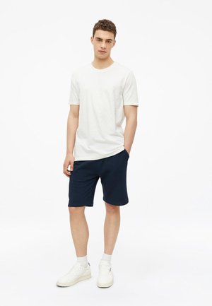 Junger Mann steht mit einer Hand in der Tasche, trägt ein weißes T-Shirt, marineblaue Shorts, weiße Socken und weiße Turnschuhe vor weißem Hintergrund.