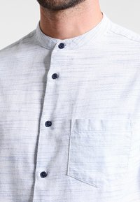 Chemise bleu clair à boutons avec col mao, en tissu texturé, rayures subtiles et boutons bleu marine. Comprend une poche poitrine.